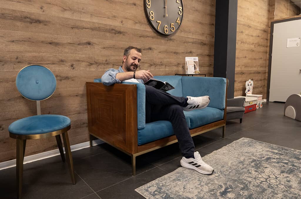 Ein Mann sitzt lesend auf einem blauen Sofa, lächelt leicht, helle Holzwand im Hintergrund.